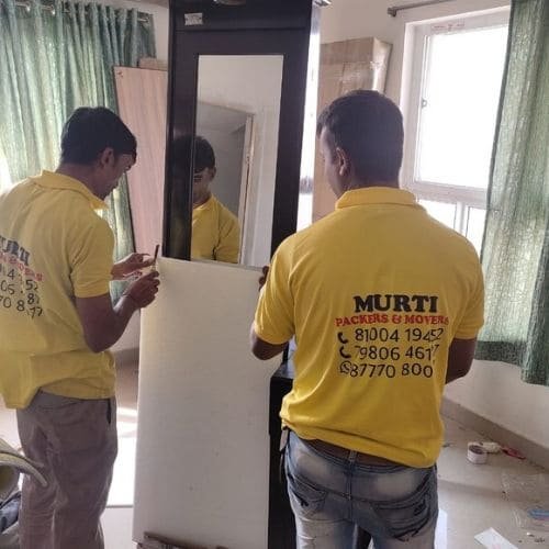 Murti packers and movers in Behala carefully wrapping a large wooden dressing table mirror with protective sheets during a home relocation service.